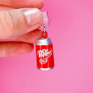 May include: Miniature red Dr. Pepper soda can charm with white text and logo. The small can has a silver bottom and a clear top loop for hanging. The charm is held against a pink background.