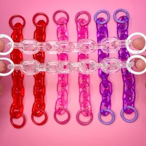 May include: Six colorful chain link accessories in red, pink, and purple. Each chain has a clear central section and a matching colored ring at each end. White circular clasps are attached to the top of each chain. The background is a solid pink.