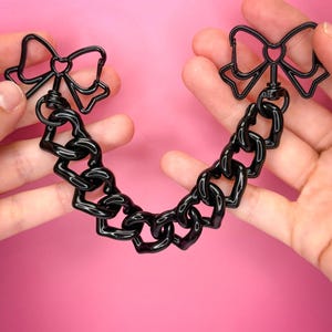 May include: A black chain accessory with bow-shaped connectors. The chain links are thick and glossy, and the bows are outlined. The accessory is held against a bright pink background.