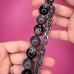 May include: A keychain featuring a thick, dark metallic chain alongside a strand of alternating beads. The beads consist of smooth black spheres and glittery dark gray spheres, with several black spider-shaped charms attached to the beads.