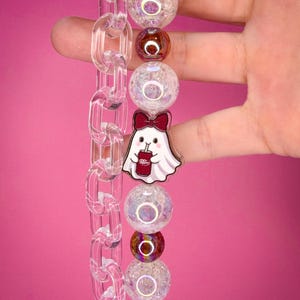 May include: A keychain featuring a clear acrylic chain link strap alongside a strand of beads. The beads include iridescent white crackle spheres and smaller metallic brown spheres. A ghost charm with a red bow holding a Dr. Pepper can is attached.