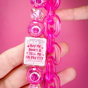 May include: A keychain featuring a rectangular charm printed with the text BUY ME BOOKS & TELL ME I'M PRETTY on a guest check design. The charm is accented with pink crackle beads, clear geometric beads, and a bright pink plastic chain link.