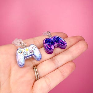 May include: Two small video game controller charms with metal loops. One charm is light blue and white with pastel buttons, while the other is dark purple with neon-style outlines and black buttons. Both feature a glossy, resin-like finish.