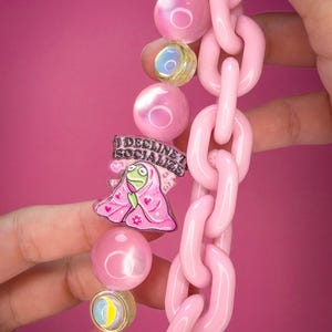 May include: A necklace featuring a thick, light pink plastic chain link strand paired with a strand of round, iridescent pink beads. An acrylic charm depicts a green frog wrapped in a pink blanket with the text I DECLINE TO SOCIALIZE.