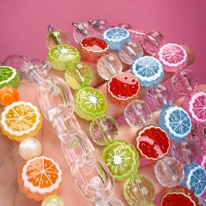May include: A collection of colorful beaded strands featuring various fruit-shaped charms including citrus slices, kiwi, and watermelon segments. The strands are interspersed with clear, faceted, and solid-colored round beads, along with clear plastic chain links.