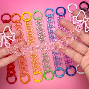 May include: A collection of colorful chain link accessories, including keychains and charms. The keychains feature a variety of colors, including red, orange, yellow, green, blue, purple, and white. The charms are shaped like bows and hearts.