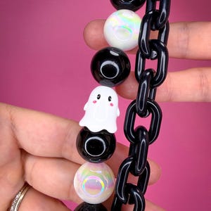 May include: A necklace featuring a strand of alternating black and iridescent white spherical beads, a white ghost charm with pink cheeks, and a thick black plastic chain link section. The design combines smooth round beads with a chunky chain.