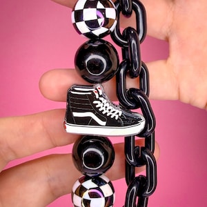May include: A necklace featuring a black and white high-top sneaker charm, alternating solid black beads, black and white checkered beads, and a thick black plastic chain link strand. The design incorporates a repeating pattern of spherical beads.