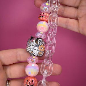 May include: A keychain featuring a series of colorful beads and a clear plastic chain. The beads include iridescent spheres, orange pumpkin-shaped charms, and a central focal piece depicting three horror movie characters surrounded by flowers.