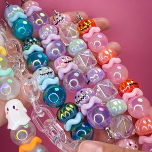 May include: A collection of beaded lanyards featuring iridescent spherical beads, clear acrylic chain links, and charms shaped like ghosts and jack-o'-lanterns. The beads are arranged in various colors including blue, purple, pink, and orange.