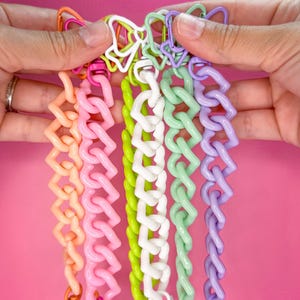 May include: A collection of colorful chain link bracelets in shades of orange, pink, white, green, and purple. The bracelets are held against a pink background, showcasing their vibrant colors and unique design. Each bracelet features a different color.