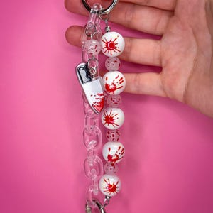 May include: A keychain featuring a silver metal ring, a clear acrylic chain, and a series of white beads. The beads are decorated with red blood splatter and handprint patterns. A silver-toned knife charm with red paint accents hangs from the chain.
