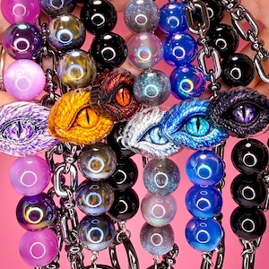 May include: A collection of five keychains featuring dark metal chains and rings. Each keychain includes a central sculpted dragon eye in various colors like purple, orange, blue, and black, surrounded by spherical beads in matching iridescent tones.