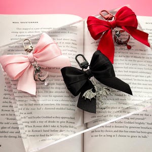 May include: Keychains with decorative bows in pink, red, and black. Each bow is attached to a clear acrylic base and features a silver-colored charm. The charms include a dragon, a crescent moon, and a heart.