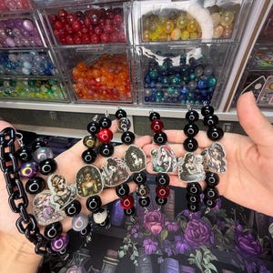 May include: A collection of beaded lanyards featuring black, red, and iridescent spherical beads interspersed with various illustrated character charms. The lanyards include a thick black plastic chain link segment and are displayed in a fan shape.