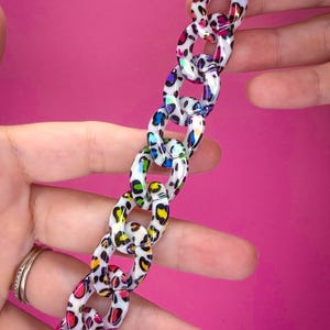 May include: A decorative chain composed of interlocking oval links featuring a white base with a multi-colored leopard print pattern in shades of pink, blue, yellow, and green. The chain is finished with a bright pink circular clasp at the top.