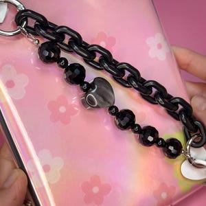 May include: Black phone charm with a chain and beaded accents. The chain is made of large, glossy links. The beaded strand features faceted black beads and a heart-shaped charm with a marbled design. The charm is attached to a pink phone case.