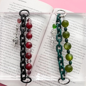May include: Two beaded chain bracelets. One is black with red beads and silver charms. The other is green with green beads and silver charms. Both are displayed on an open book.