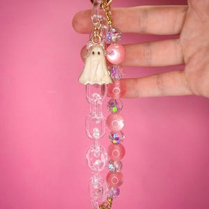 May include: A handmade keychain featuring a white ghost charm with a pink bow, clear chain links, and pink and iridescent beads. The keychain has a gold-colored clasp and ring.