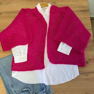Könnte beinhalten: Ein leuchtend pinkfarbener, offener Strickcardigan mit weiten Ärmeln liegt über einem weißen Button-Down-Hemd. Hellblaue Jeans sind darunter gefaltet. Die Kleidung wird auf einer Holzoberfläche präsentiert.