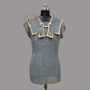 May include: A chainmail shirt with a decorative collar and shoulder accents. The shirt is displayed on a mannequin. The collar and shoulder accents are trimmed with a light brown material and secured with gold-colored clasps.