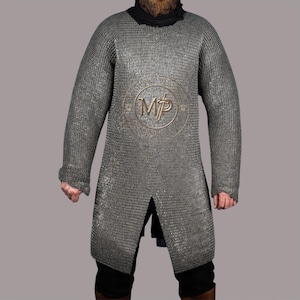 Puede incluir: Una cota de malla plateada con mangas largas y abertura frontal. La camisa tiene un emblema redondo en el centro. La persona lleva un cuello alto negro y pantalones negros. La cota de malla está hecha de pequeños anillos metálicos entrelazados.