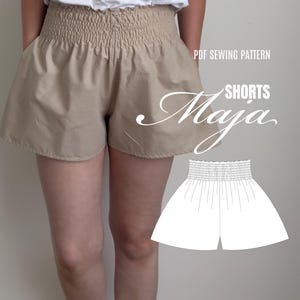 Puede incluir: Pantalones cortos de color beige con una cintura elástica fruncida ancha y un corte holgado. La imagen incluye el texto PDF SEWING PATTERN SHORTS Maja y una ilustración de línea blanca del diseño, junto con el texto the sewcial club.