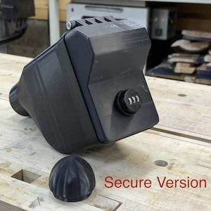 Puede incluir: Un contenedor negro rectangular con cerradura de combinaci&oacute;n y tapa redondeada extra&iacute;ble. El contenedor tiene una superficie texturizada y una extensi&oacute;n cil&iacute;ndrica en un lado. El texto "Secure Version" est&aacute; impreso en rojo.