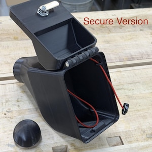 Puede incluir: Contenedor rectangular negro con tapa con bisagras y cerradura peque&ntilde;a. La tapa est&aacute; abierta, revelando un cable rojo en el interior. Una perilla negra y redondeada est&aacute; junto al contenedor. El texto en la imagen dice "Secure Version".