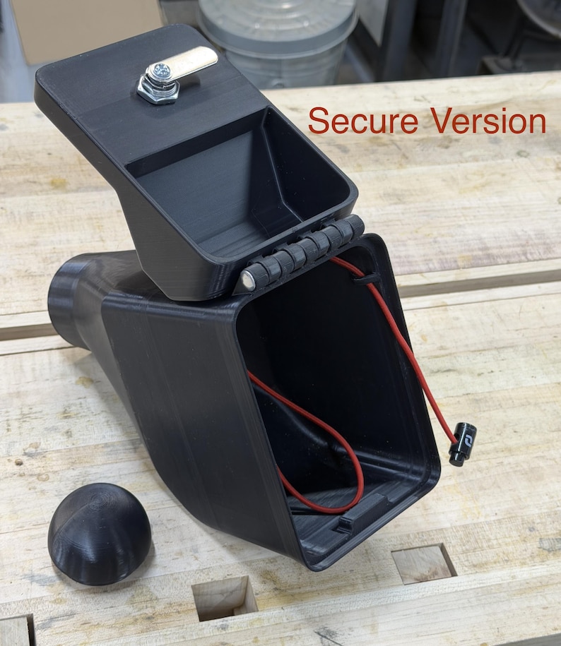 Puede incluir: Contenedor rectangular negro con tapa con bisagras y cerradura peque&ntilde;a. La tapa est&aacute; abierta, revelando un cable rojo en el interior. Una perilla negra y redondeada est&aacute; junto al contenedor. El texto en la imagen dice "Secure Version".