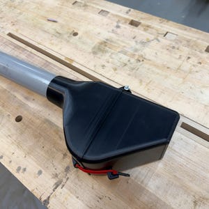 Puede incluir: Un accesorio de herramienta de pl&aacute;stico negro con un poste de metal gris. El accesorio tiene forma rectangular con una parte superior curva y un cord&oacute;n rojo. La herramienta est&aacute; sobre una superficie de madera marr&oacute;n claro.