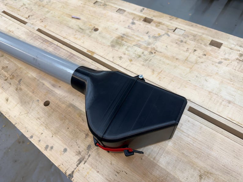 Puede incluir: Un accesorio de herramienta de pl&aacute;stico negro con un poste de metal gris. El accesorio tiene forma rectangular con una parte superior curva y un cord&oacute;n rojo. La herramienta est&aacute; sobre una superficie de madera marr&oacute;n claro.