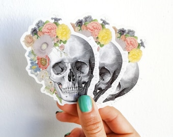 May include: Three anatomical skull stickers with floral wreaths. The skulls are black and white and the wreaths are made of pink, yellow, and green flowers.