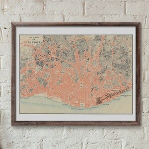 May include: A framed vintage map of Lisbon, Portugal, with the title "Plano de Lisboa" printed in the top left corner. The map is in shades of pink, blue, and green, and is displayed on a white brick wall above a wooden desk with a laptop, camera, and other office supplies.