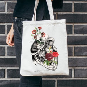 Op de afbeelding: Een witte tote bag met een zwart-witte anatomische hartillustratie en rode bloemen. De tas heeft een zwarte handvat en wordt door een persoon vastgehouden.