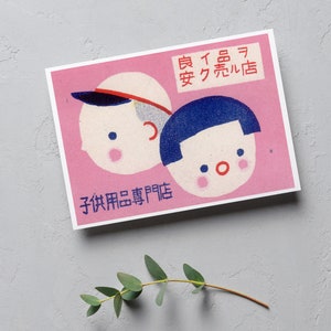 May include: A pink and white vintage matchbox with a red and blue illustration of two children's faces. The text on the matchbox reads "子供用品専門店" and "真下呉服店".