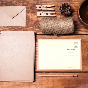 May include: A brown notebook with a spiral binding, a blank postcard, a roll of twine, a pencil, two clothespins, a pine cone, and a brown envelope on a wooden surface.
