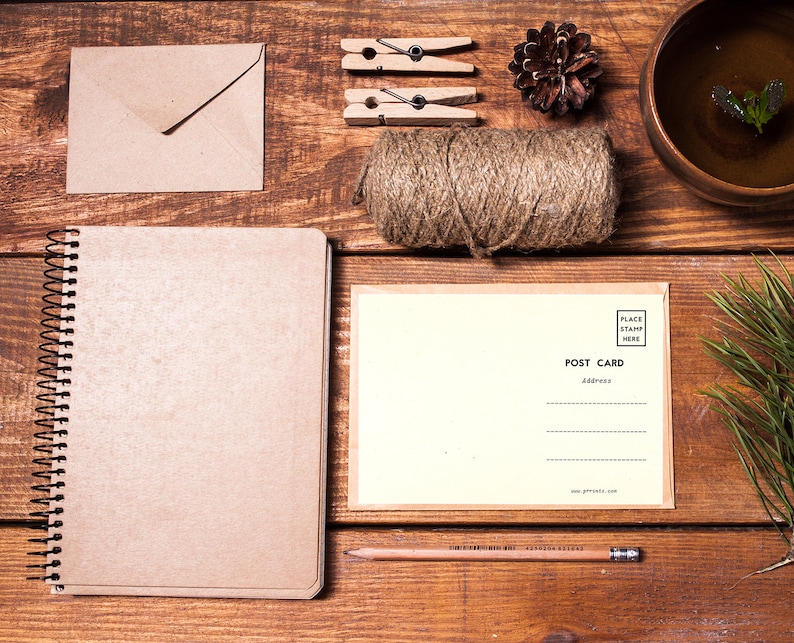May include: A brown notebook with a spiral binding, a blank postcard, a roll of twine, a pencil, two clothespins, a pine cone, and a brown envelope on a wooden surface.