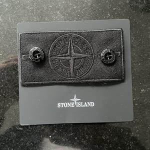 May include: A black patch featuring the Stone Island logo, with two black buttons on either side. The patch is presented on a grey card, with the Stone Island logo printed below.