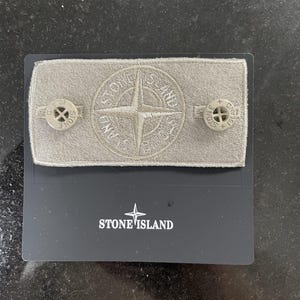 Può includere: Un distintivo Stone Island e due bottoni su una carta nera. Il distintivo è rettangolare, con una base in tessuto grigio e un logo a rosa dei venti cucito. I bottoni circolari presentano un disegno a croce.