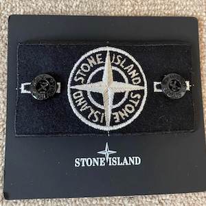 Op de afbeelding: Een zwart rechthoekig embleem met de woorden "STONE ISLAND" en een wit geborduurd kompasrooslogo. Twee zwarte knopen zijn aan het embleem bevestigd. Het embleem bevindt zich op een zwarte kaart.