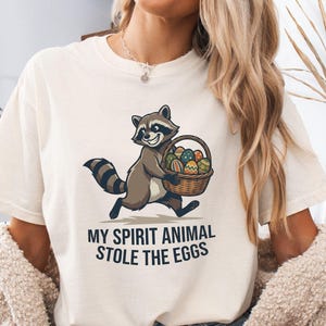 Op de afbeelding: Crèmekleurig T-shirt met een cartoon wasbeer die een mand met paaseieren draagt. De tekst "MY SPIRIT ANIMAL STOLE THE EGGS" staat onder de wasbeerillustratie. Het T-shirt is een casual unisex stijl.