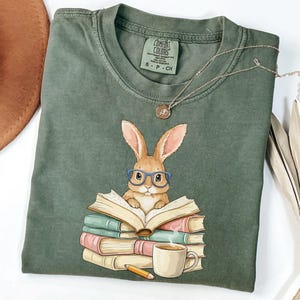 May include: Sage green t-shirt featuring a cartoon rabbit wearing glasses and reading a stack of books. A cup of coffee and a pencil are next to the books. The shirt has a Comfort Colors tag.