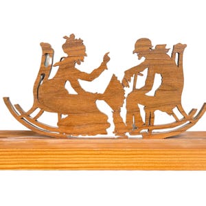 May include: A wooden silhouette depicting two figures seated in rocking chairs, engaged in a conversation. The figures are carved from a light brown wood and mounted on a rectangular wooden base. The piece is a decorative item.