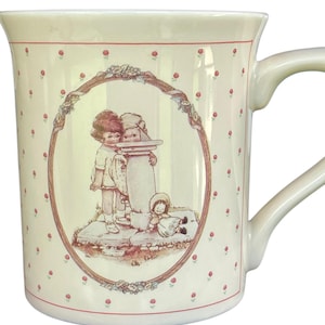 May include: A cream-colored ceramic mug featuring a repeating pattern of small red rosebuds. The center displays an oval illustration of two children standing by a sundial with a small doll sitting on the ground nearby, all framed by a floral border.