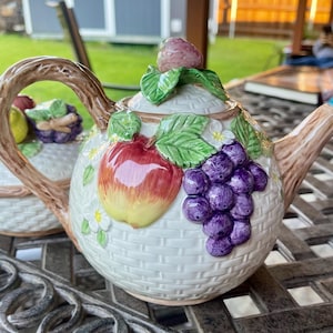 May include: A ceramic teapot with a basket weave design, adorned with a red apple, purple grapes, and green leaves. The handle and spout have a branch-like appearance. The lid is topped with a strawberry and more leaves. The teapot is white with colorful accents.