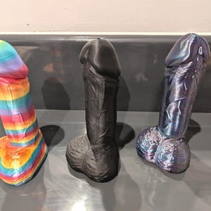 May include: Three 3D-printed adult toys. One is rainbow-coloured, the second is solid black, and the third is a dark blue with a metallic sheen. All three are standing upright on a grey surface.