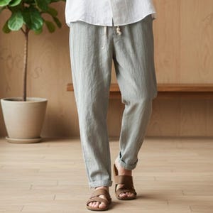 May include: A person wearing a white short-sleeved shirt and gray linen pants. The shirt has a mandarin collar and button-down front. The pants have a drawstring waist. The person is wearing brown sandals. The background is a wooden wall and floor.