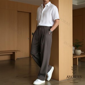 May include: A person wearing a white short-sleeve shirt and dark brown pleated trousers. The person is also wearing a watch and white sneakers. The image includes the text "AYORAS LINEN & COTTON CLOTHING SHOP WOMEN AND MEN".