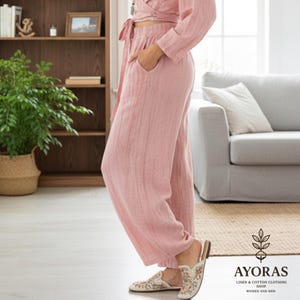 May include: A light pink linen outfit, including a wrap top and wide-leg pants. The outfit has a relaxed fit and features pockets. The person is wearing floral embroidered slip-on shoes. The image is taken indoors, with a couch and bookshelf in the background.